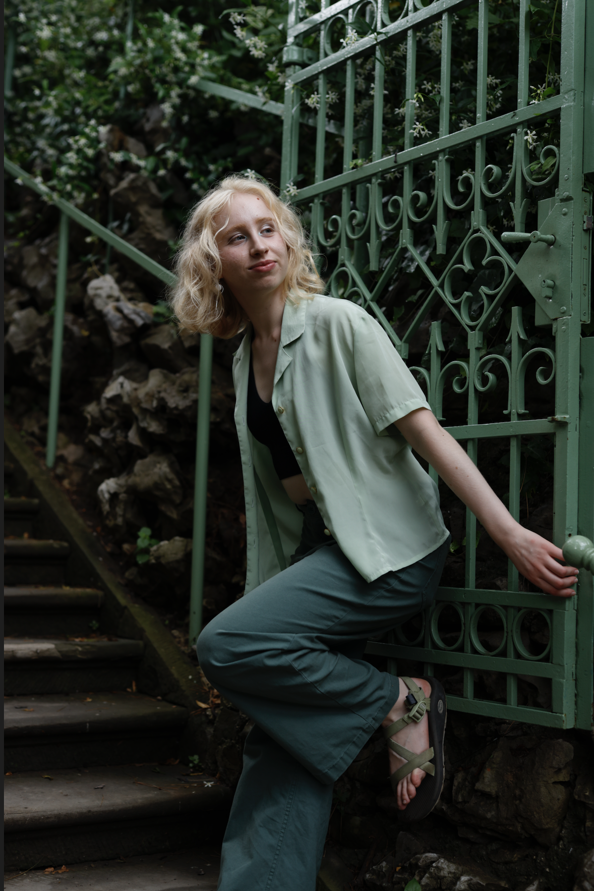 A picture of Hannah as she peaks around the corner of a garden gate. One of her feet is proped up on the gate and her hands anchor her as she leans away. Her dark sage pants and mint shirt match the green of the metal and jasmine behind her.  She has a slight smile, a look of mischief.