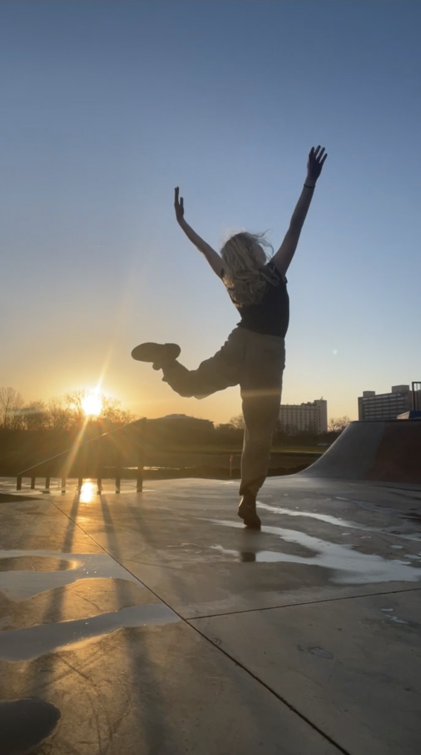 A picture of Hannah dances on a wet skate park. Its golden hour and the sun reflects off of puddles. Hannah is in the middle of tuning, balanced on one leg, with her arms reaching up.