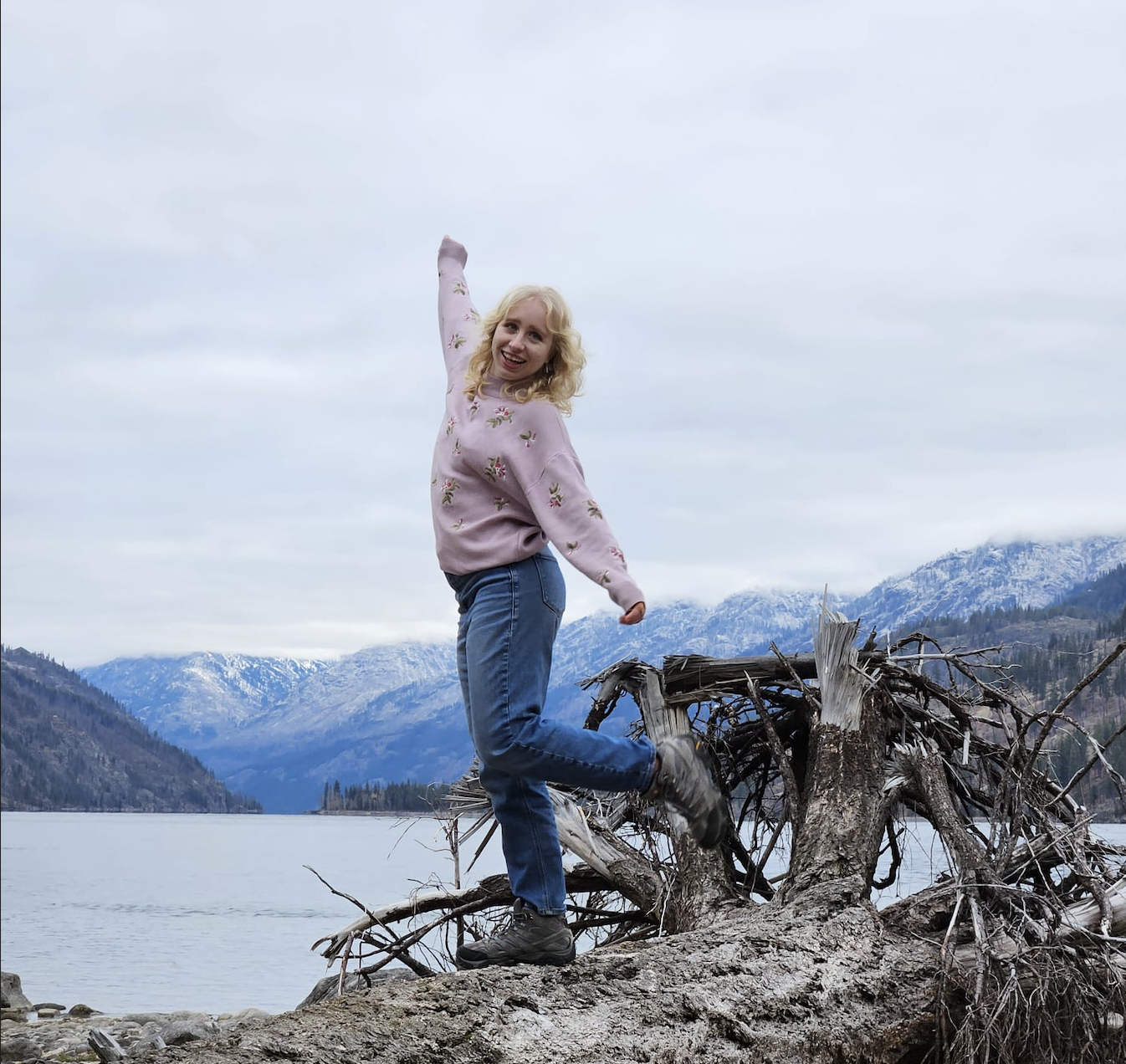 A picture of Hannah as she balances on a fallen tree on the beach of a lake. She stares off into the distance playfully, with her front leg lifted at the knee. In the background, there are icy blue mountains that surround the lake.